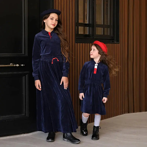 Two children in matching navy blue dresses with red accents standing in front of a wooden paneled wall.